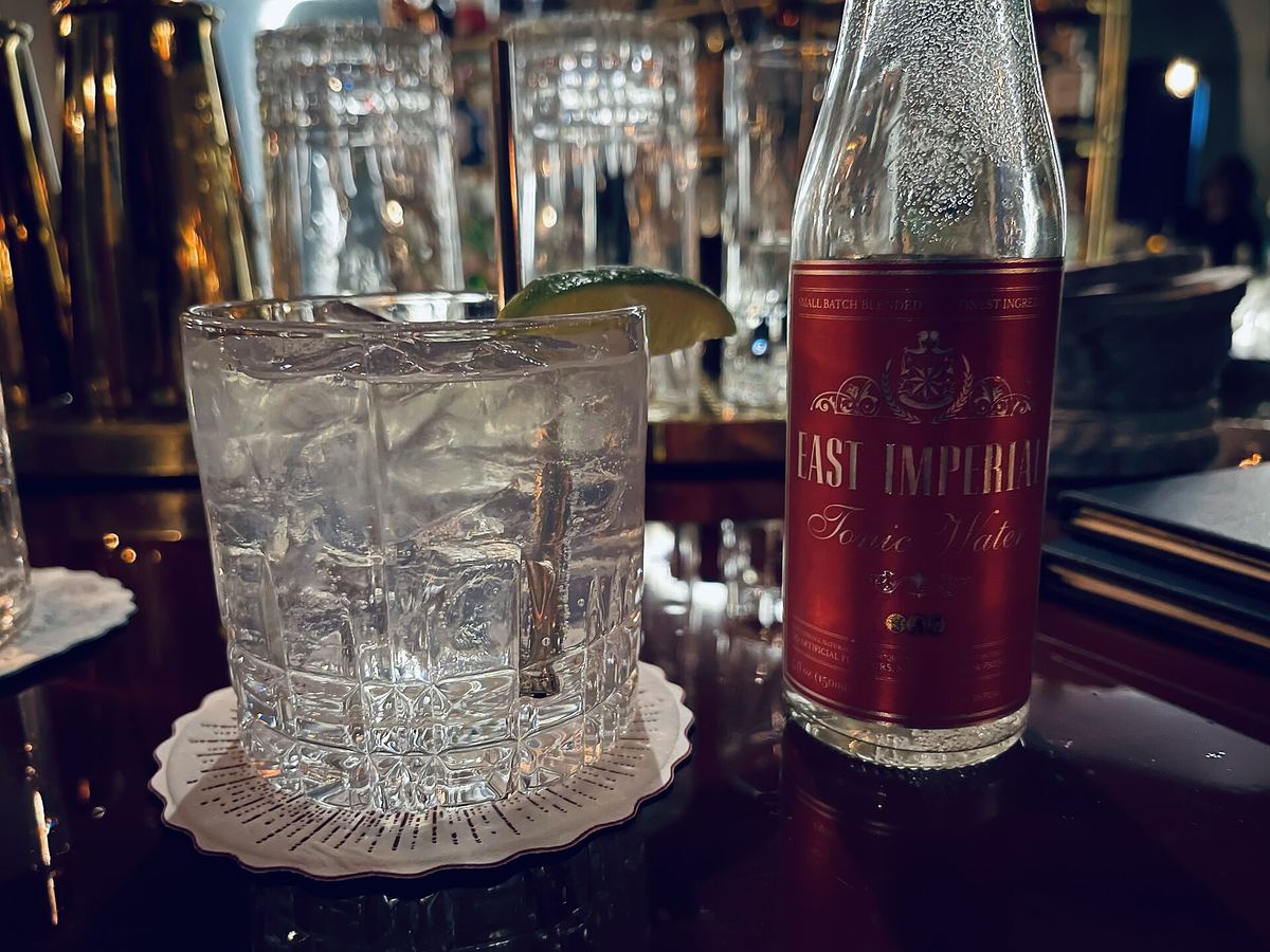 Gin tonic con lime su un bancone in legno scuro, accanto a una bottiglia di acqua tonica